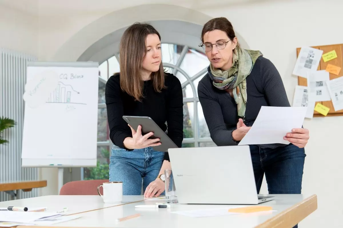 Zwei Menschen blicken auf einen Laptop und besprechen etwas. Eine Person hat ein Tablet in der Hand, die andere ein Blatt Papier.